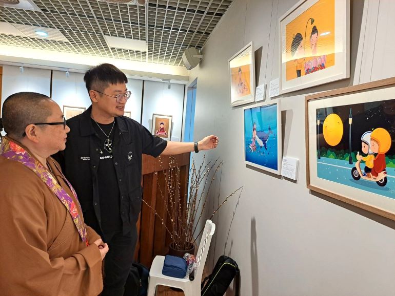 Q版佛畫映照慈悲世界 丁丁個展三重禪淨中心溫暖登場 21 《圖說》張丁文為三重禪淨中心監寺如宗法師(左)介紹「聲聲阿彌陀佛」(圖右下)的創作緣由。〈佛光山三重禪淨中心提供〉